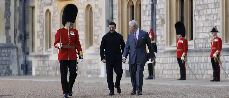 Two men, one in a suit and the other in a black coat, walk together outside a historic stone building.