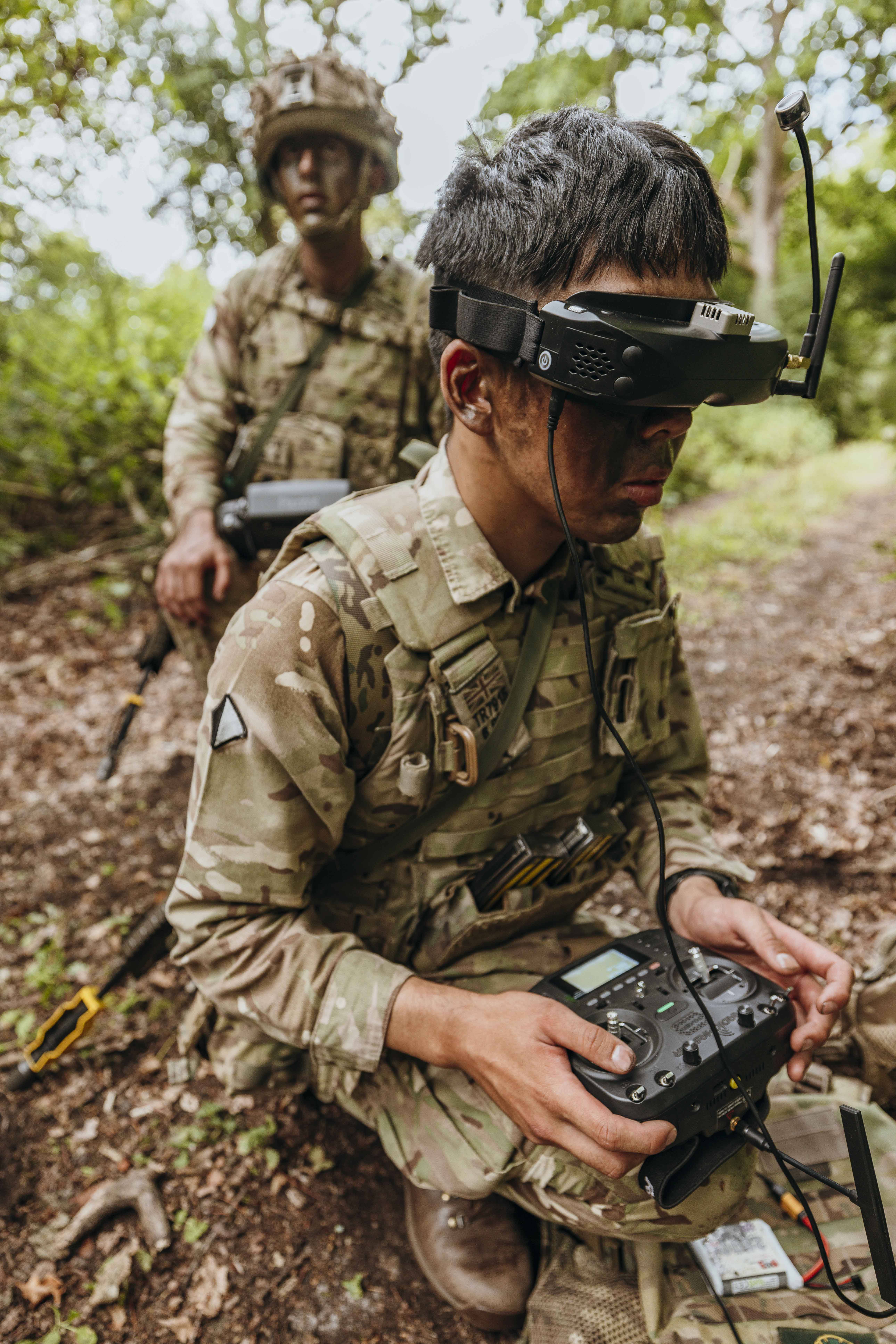 Soldier in camouflage gear operating a remote control device with antenna in a wooded area during a military exercise.