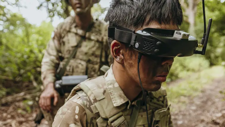 Soldier in camouflage gear operating a remote control device with antenna in a wooded area during a military exercise.
