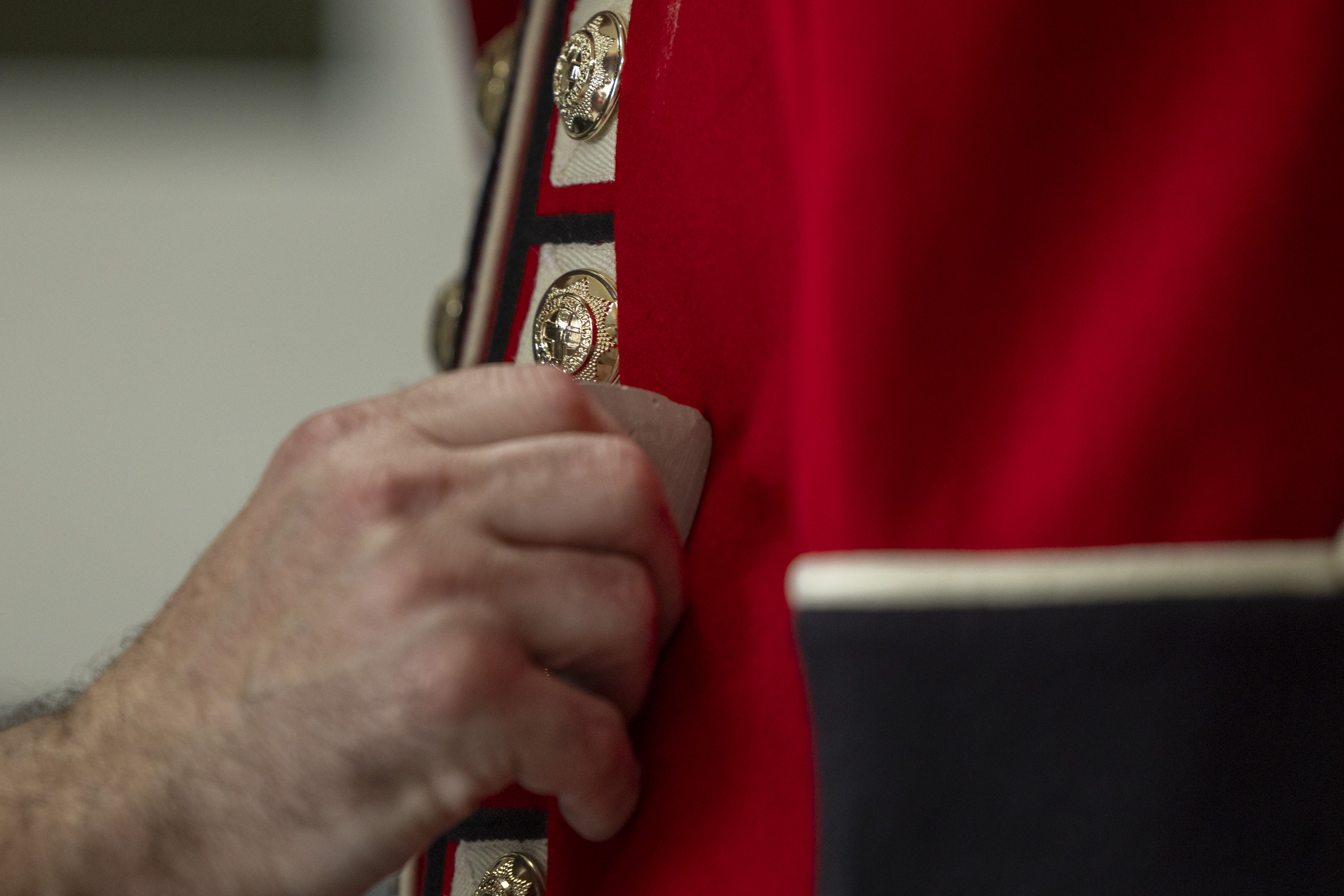 This image shows someone buttoning a red tunic as the ceremonial dress of the Household Division. The jacket features coldstream guards cap badge imprinted on shiny, metallic buttons going down the front of the tunic.