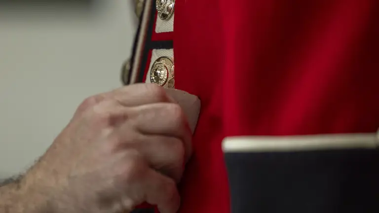 This image shows someone buttoning a red tunic as the ceremonial dress of the Household Division. The jacket features coldstream guards cap badge imprinted on shiny, metallic buttons going down the front of the tunic.