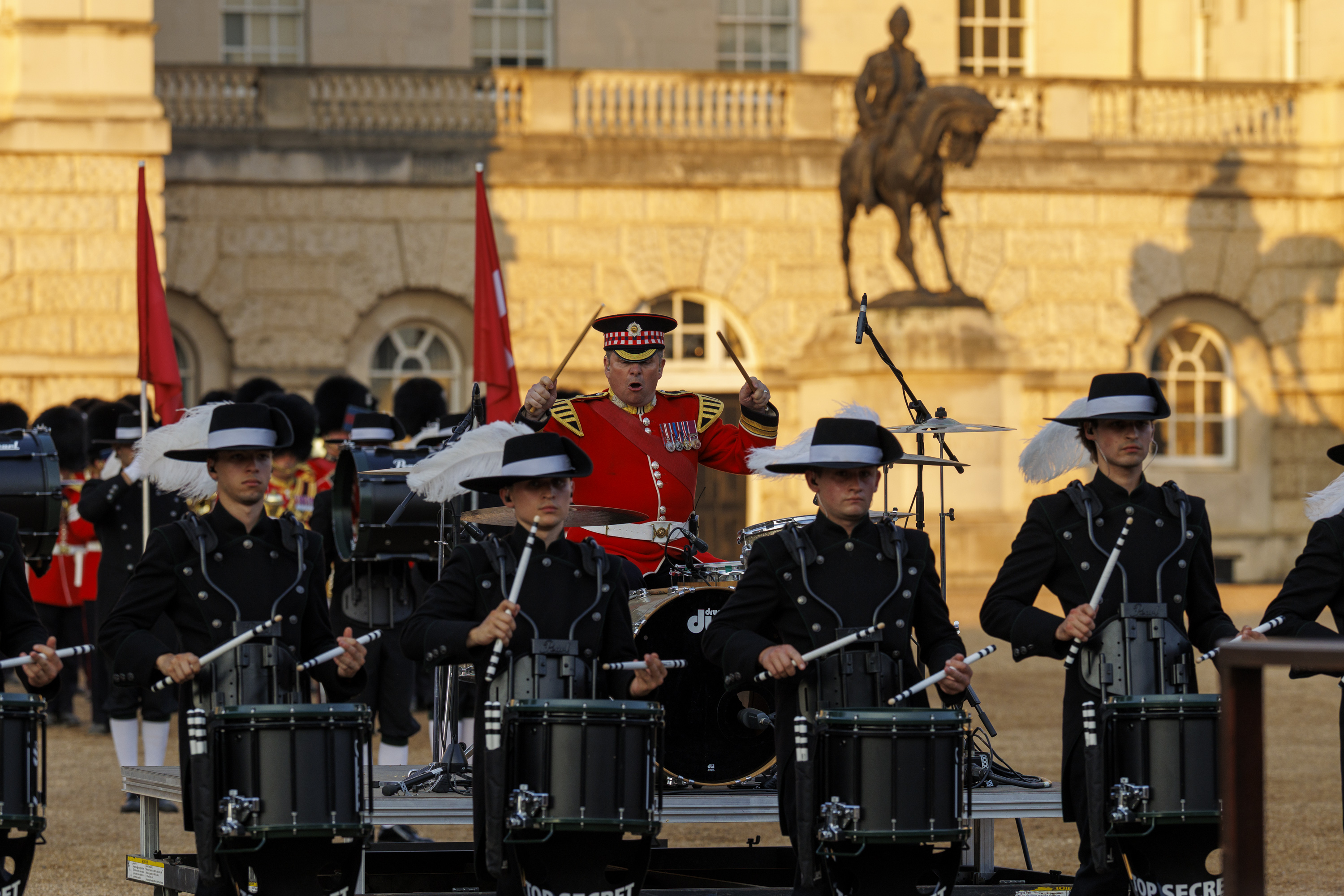 A drum corps in black uniforms performs in front of a historic building. Behind them, a figure in a red uniform stands with a drum. The backdrop includes an equestrian statue and flags.