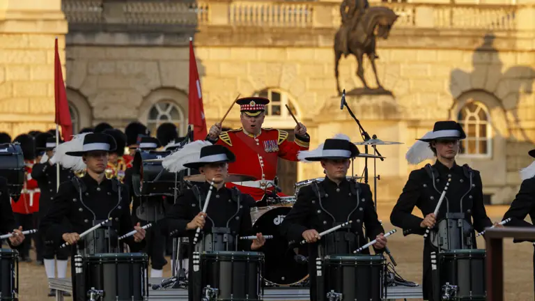 A drum corps in black uniforms performs in front of a historic building. Behind them, a figure in a red uniform stands with a drum. The backdrop includes an equestrian statue and flags.