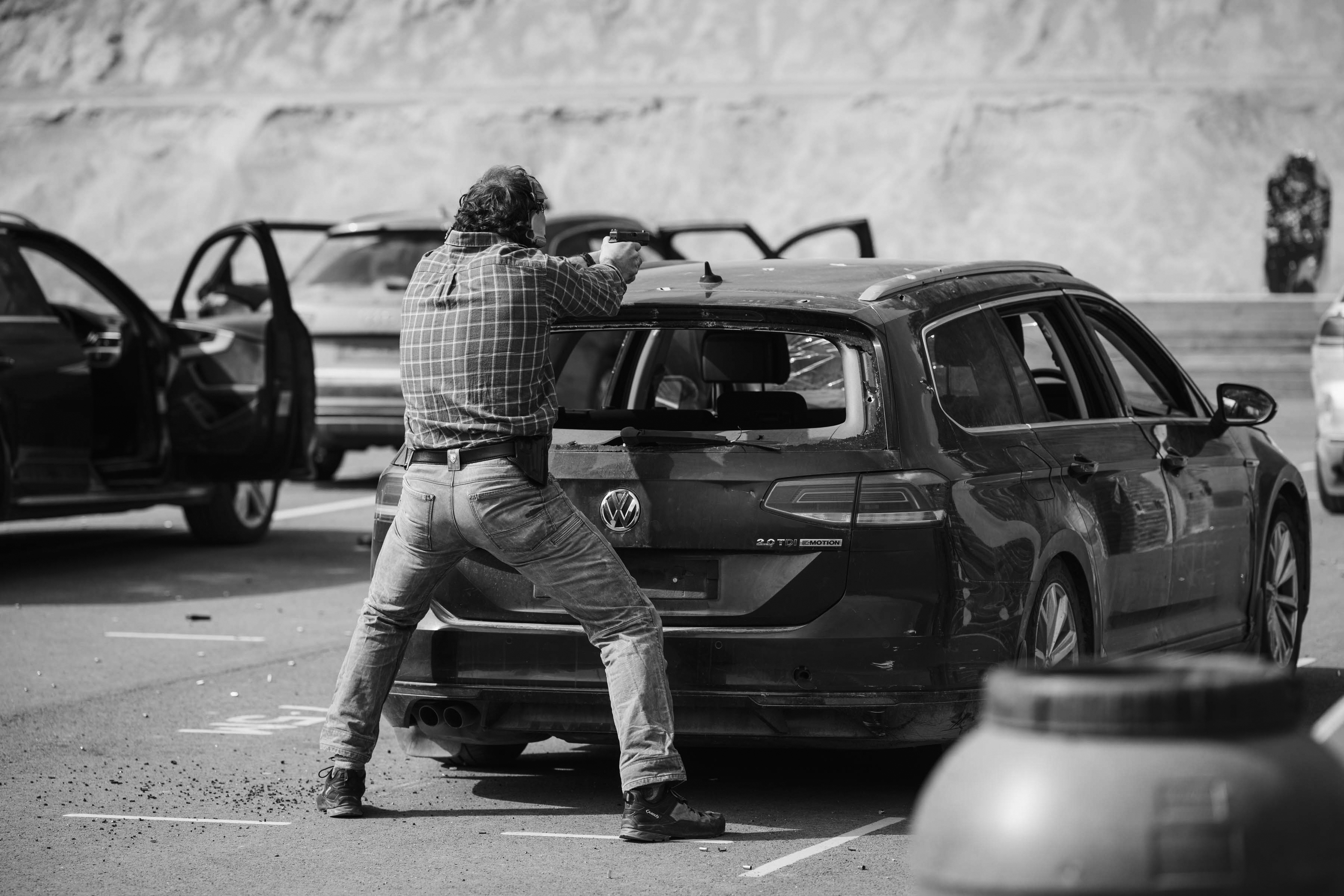A person aims a pistol through the rear window of a parked Volkswagen car in a parking lot. Other cars are nearby.