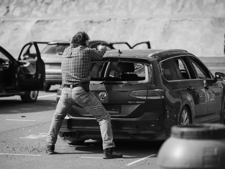 A person aims a pistol through the rear window of a parked Volkswagen car in a parking lot. Other cars are nearby.