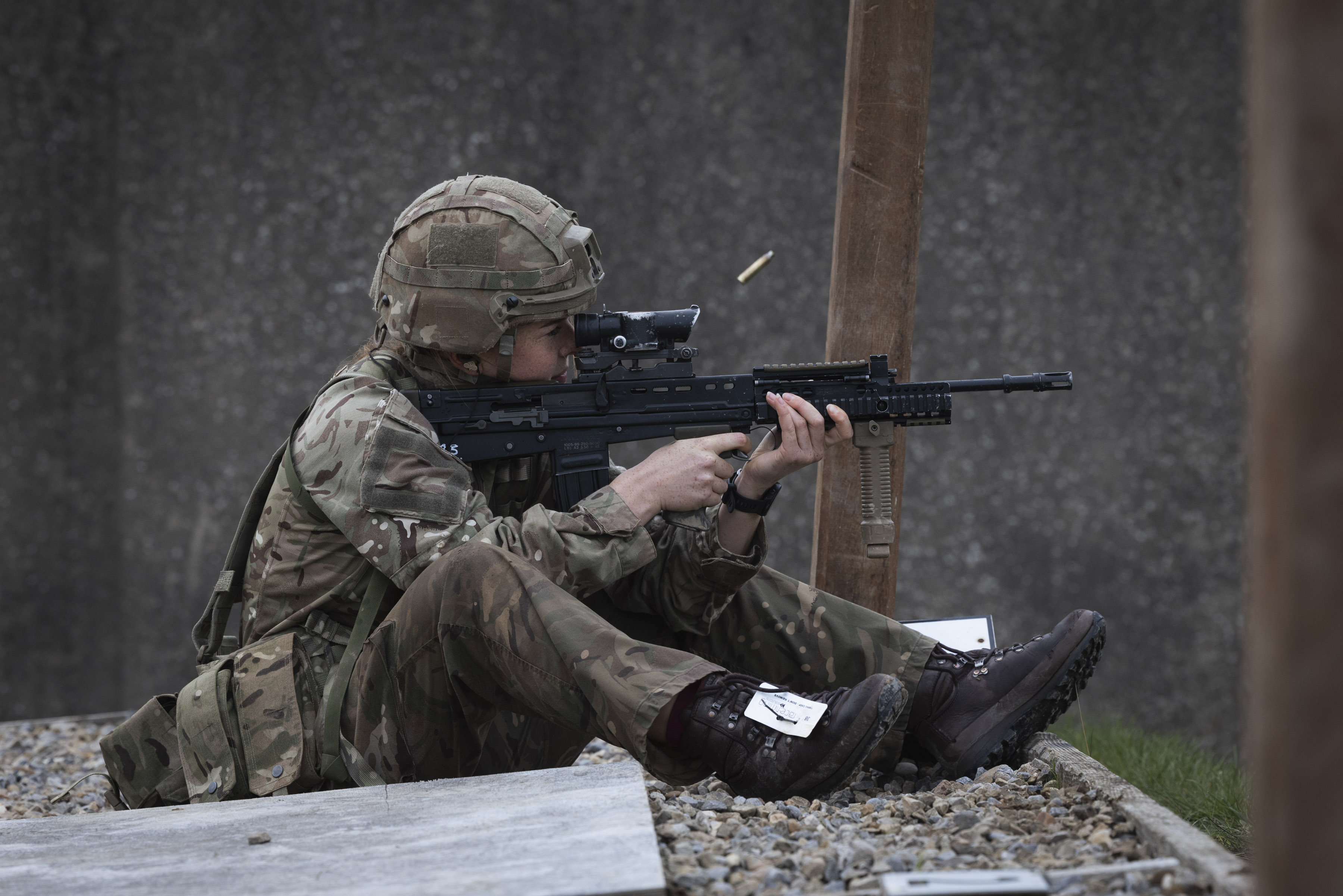 Soldier in full camouflage gear sitting on gravel, aiming a rifle with a scope during a training exercise.