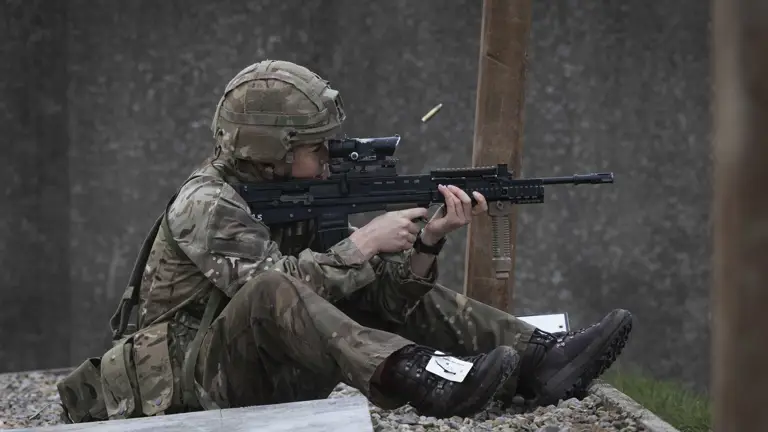 Soldier in full camouflage gear sitting on gravel, aiming a rifle with a scope during a training exercise.