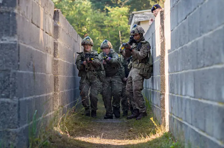 A group of soldiers in camouflage clothing and helmets move cautiously through a narrow, walled passageway. They are holding rifles and appear focused on their surroundings. In the background, vegetation is visible beyond the walls.