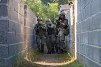 A group of soldiers in camouflage clothing and helmets move cautiously through a narrow, walled passageway. They are holding rifles and appear focused on their surroundings. In the background, vegetation is visible beyond the walls.