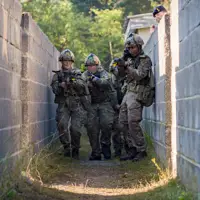 A group of soldiers in camouflage clothing and helmets move cautiously through a narrow, walled passageway. They are holding rifles and appear focused on their surroundings. In the background, vegetation is visible beyond the walls.
