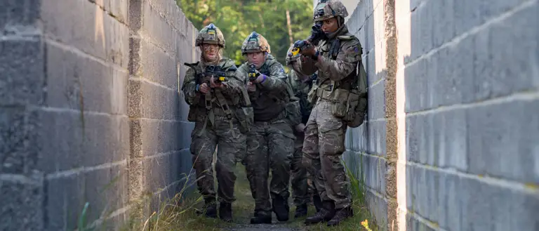 A group of soldiers in camouflage clothing and helmets move cautiously through a narrow, walled passageway. They are holding rifles and appear focused on their surroundings. In the background, vegetation is visible beyond the walls.
