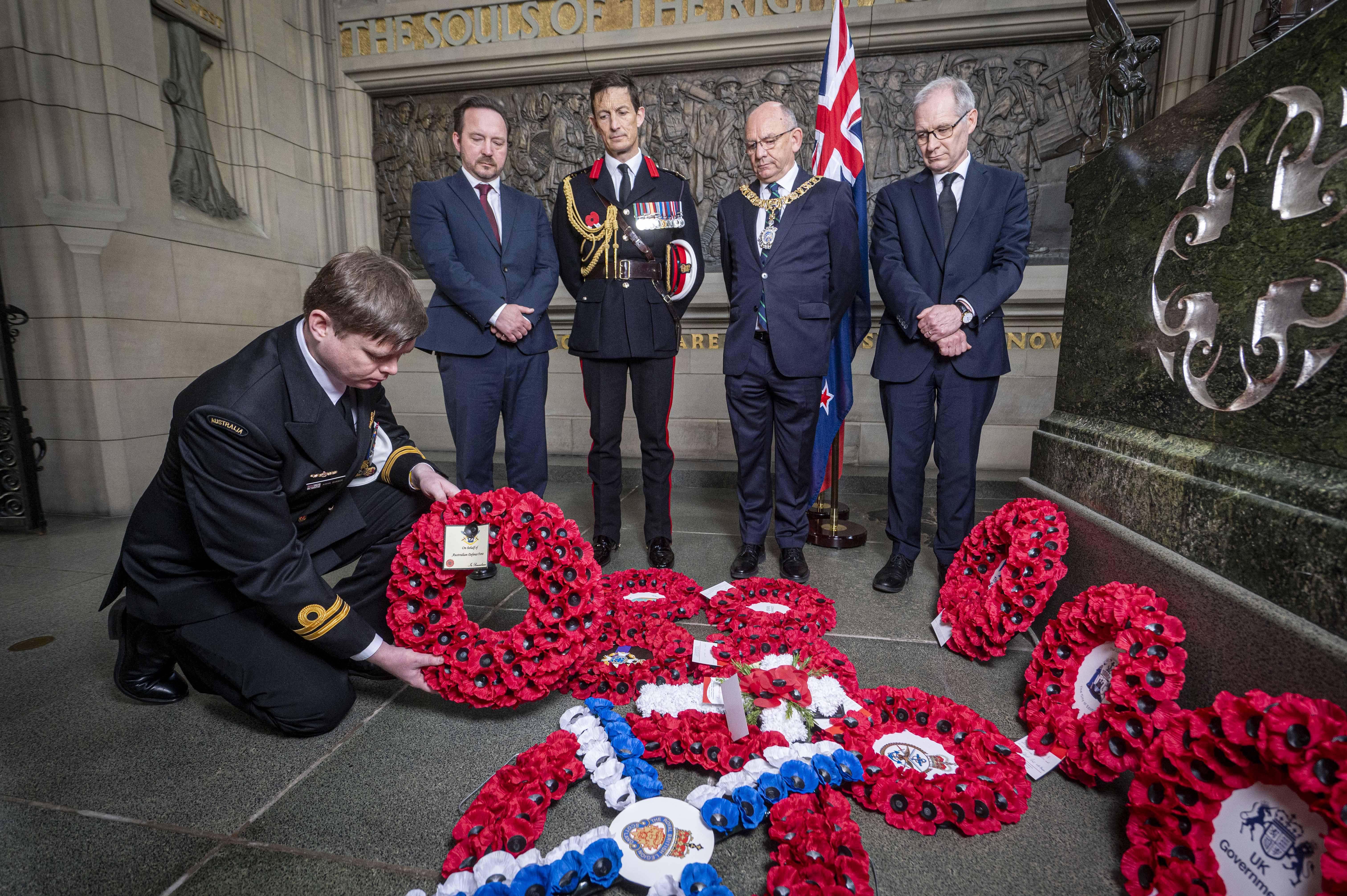 An Australian soldier in black ceremonial uniform lays a wreath in front of four others.
