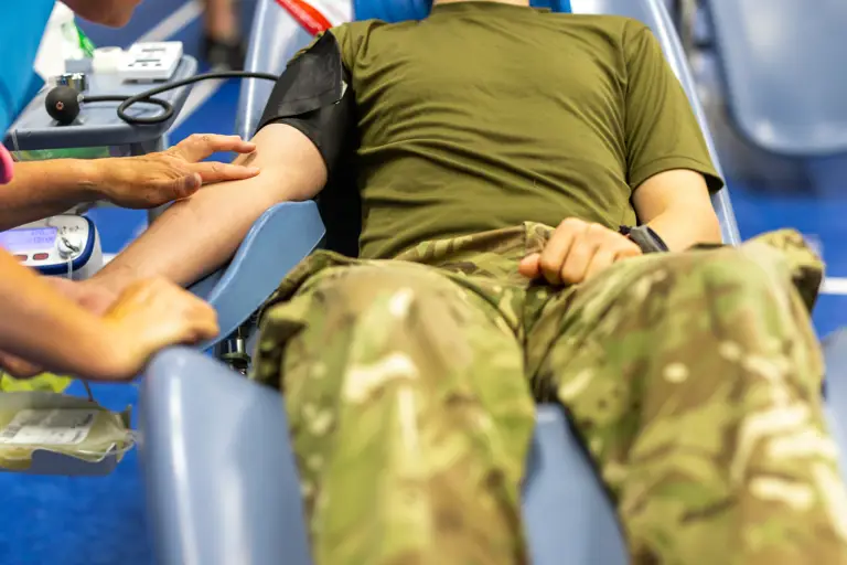 A soldier in camouflage trousers and olive green shirt donates blood while seated in a medical chair during a blood drive.