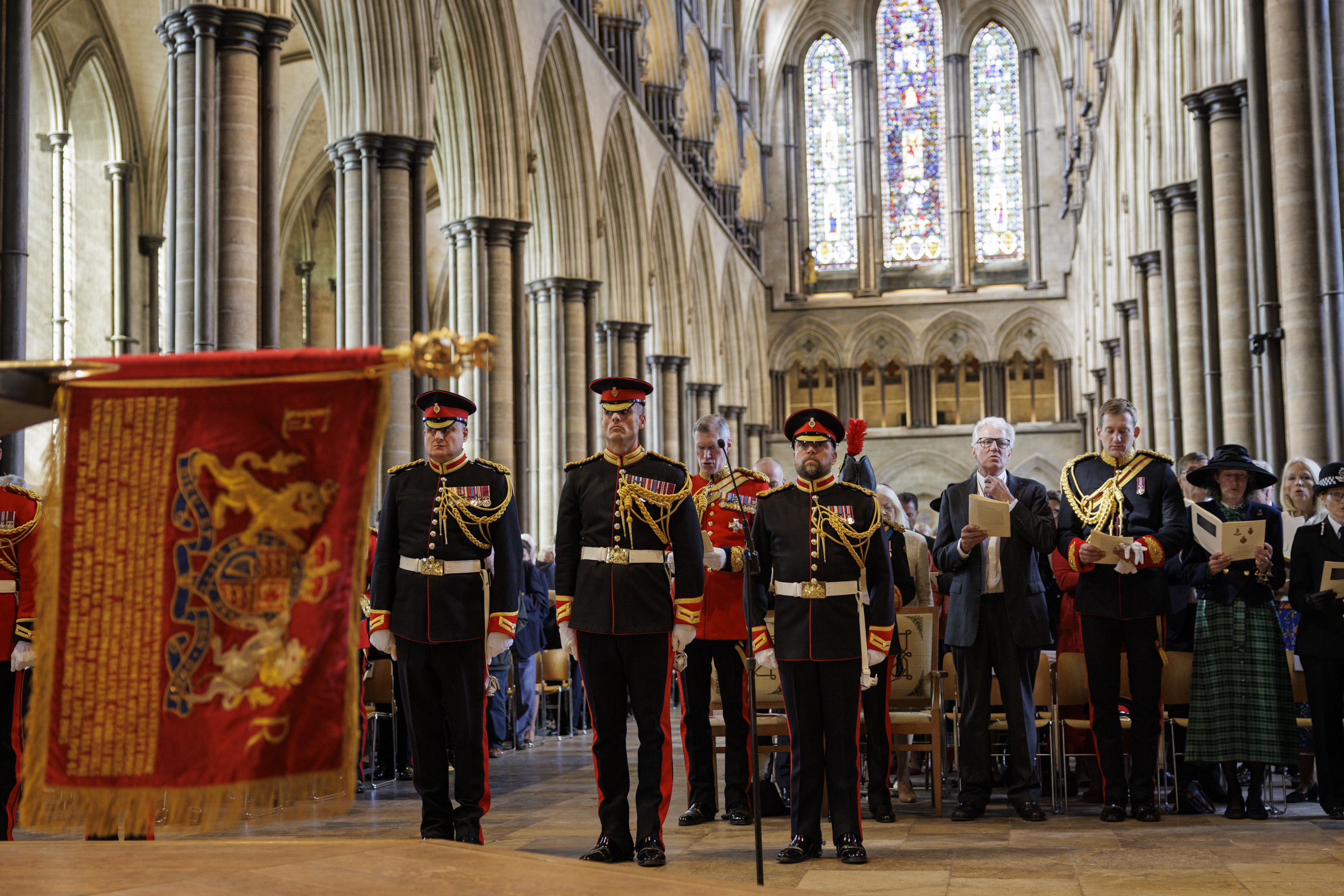 Men in black ceremonial uniforms stand in front of a congregation. A red and gold ceremonial flag is laid on a stone baptismal font.