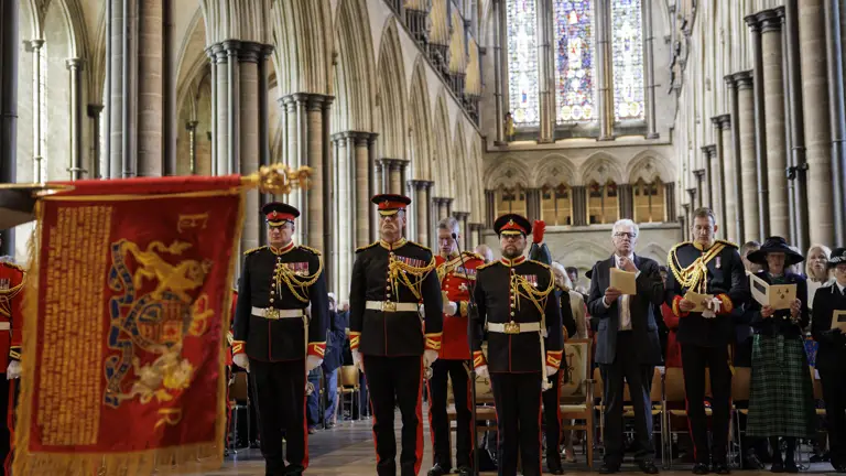 Men in black ceremonial uniforms stand in front of a congregation. A red and gold ceremonial flag is laid on a stone baptismal font.