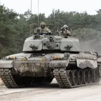 A military tank, the Challenger 2, is on a dirt road surrounded by a forested area. Two individuals are visible on the tank.