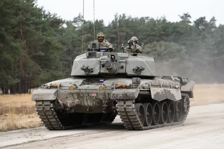 A military tank, the Challenger 2, is on a dirt road surrounded by a forested area. Two individuals are visible on the tank.