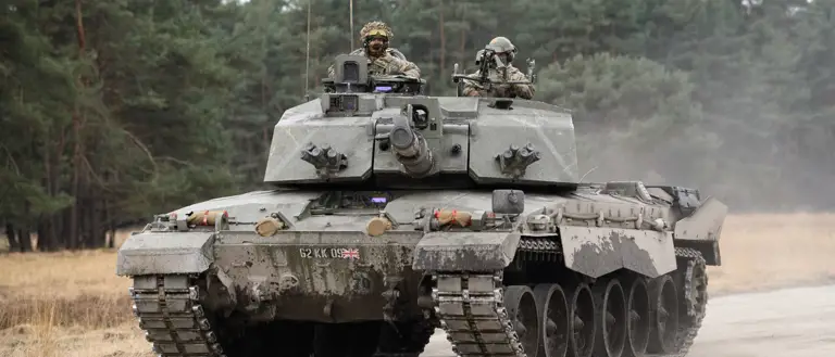 A military tank, the Challenger 2, is on a dirt road surrounded by a forested area. Two individuals are visible on the tank.