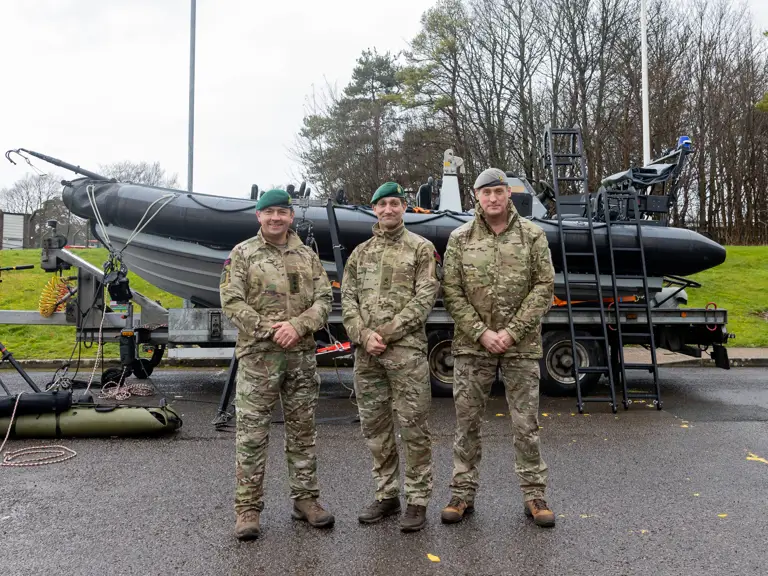 Three soldiers in camouflage uniforms stand in front of a black inflatable military boat on a trailer in an outdoor setting.