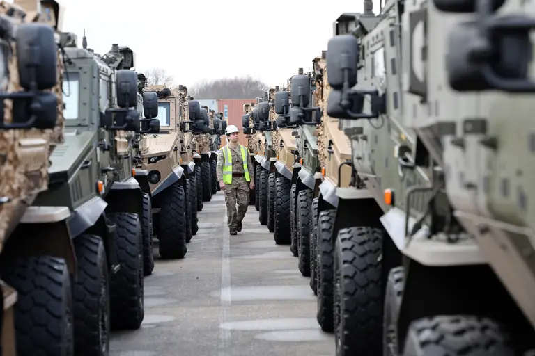 Soldier in camouflage uniform is seen walking between lined up vehicles while wearing a hi vis jacket and a hard hat checking vehicles over before transportation.