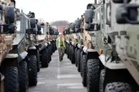 Soldier in camouflage uniform is seen walking between lined up vehicles while wearing a hi vis jacket and a hard hat checking vehicles over before transportation.
