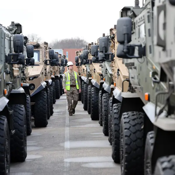 Soldier in camouflage uniform is seen walking between lined up vehicles while wearing a hi vis jacket and a hard hat checking vehicles over before transportation.