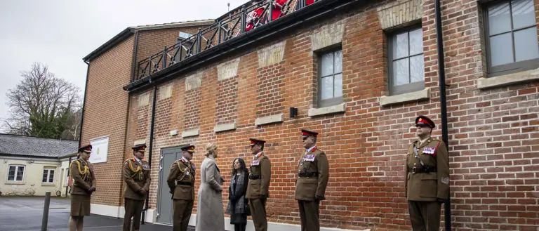 Her Royal Highness The Duchess of Edinburgh is greeted soldiers in khaki uniforms. Behind them is a brick built building
