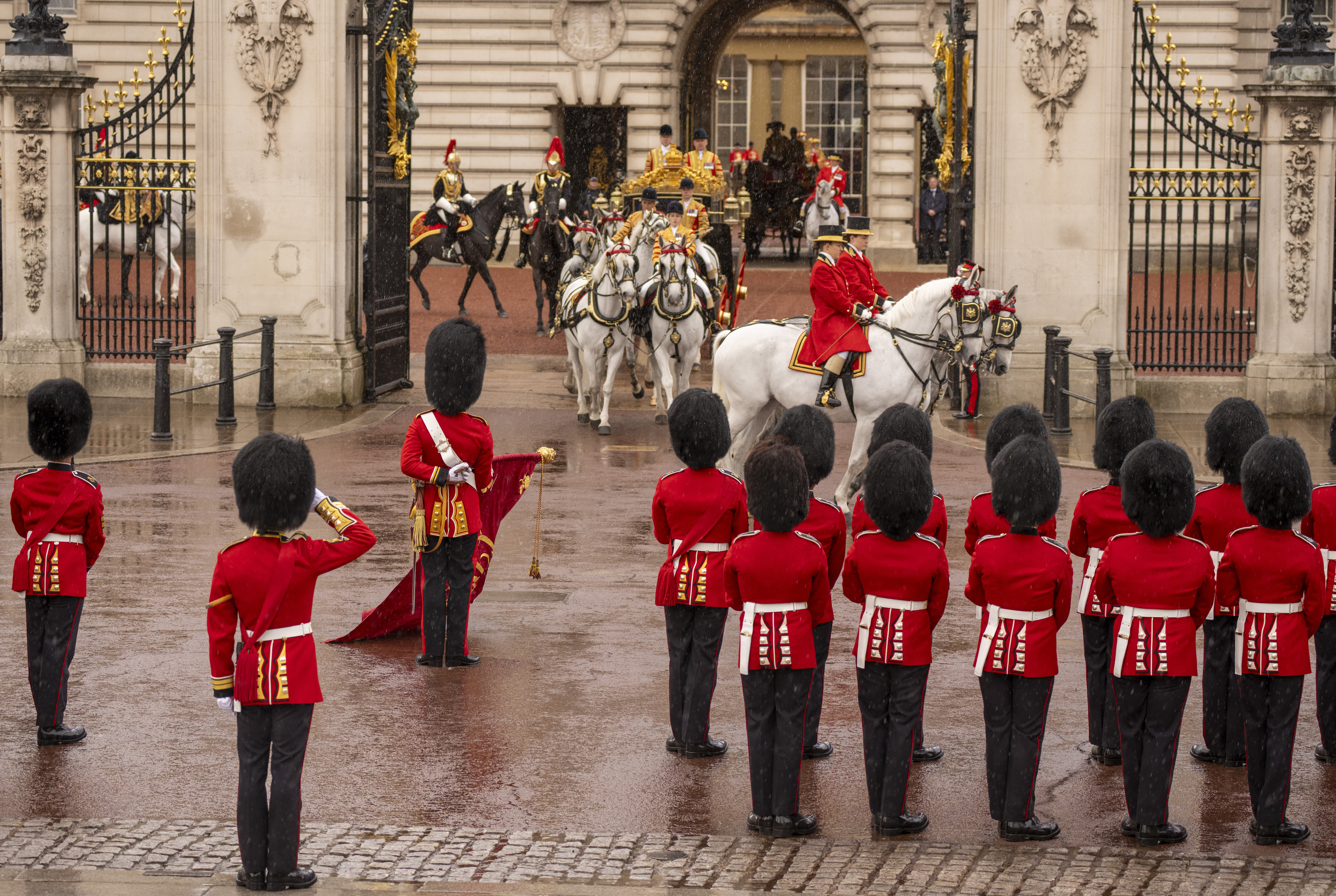 A group of ceremonial guards in red uniforms with tall black hats stands in formation outside Buckingham Palace. 