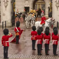A group of ceremonial guards in red uniforms with tall black hats stands in formation outside Buckingham Palace.