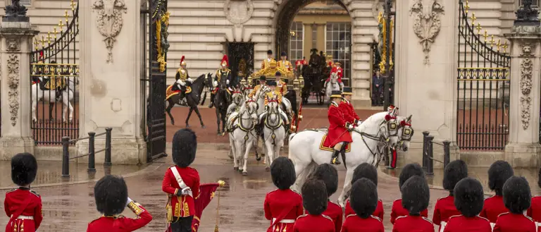 A group of ceremonial guards in red uniforms with tall black hats stands in formation outside Buckingham Palace.