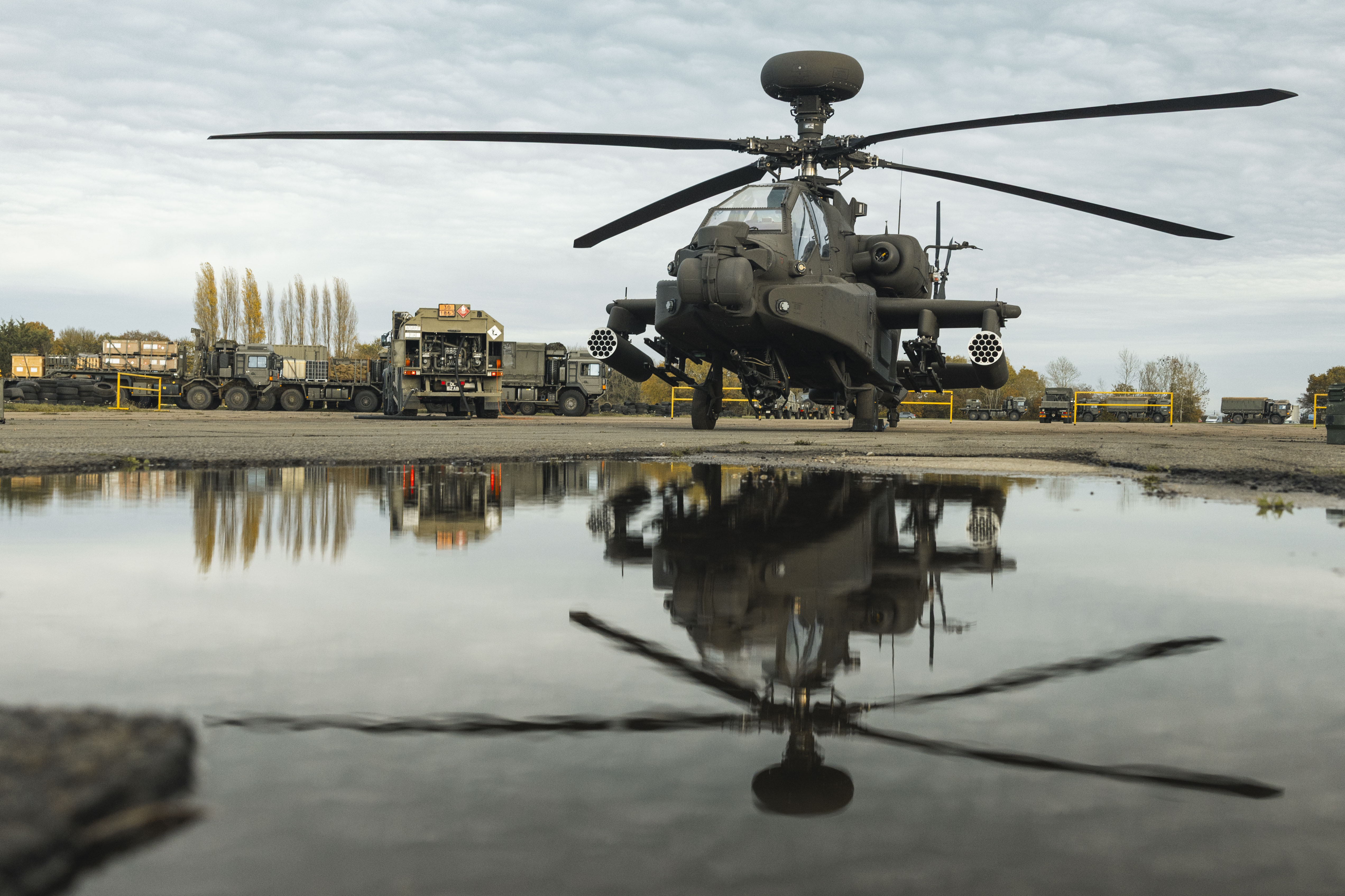 Military attack helicopter parked on a tarmac with its reflection visible in a nearby puddle under a cloudy sky.