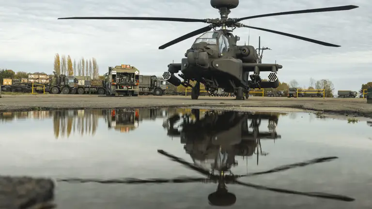 Military attack helicopter parked on a tarmac with its reflection visible in a nearby puddle under a cloudy sky.