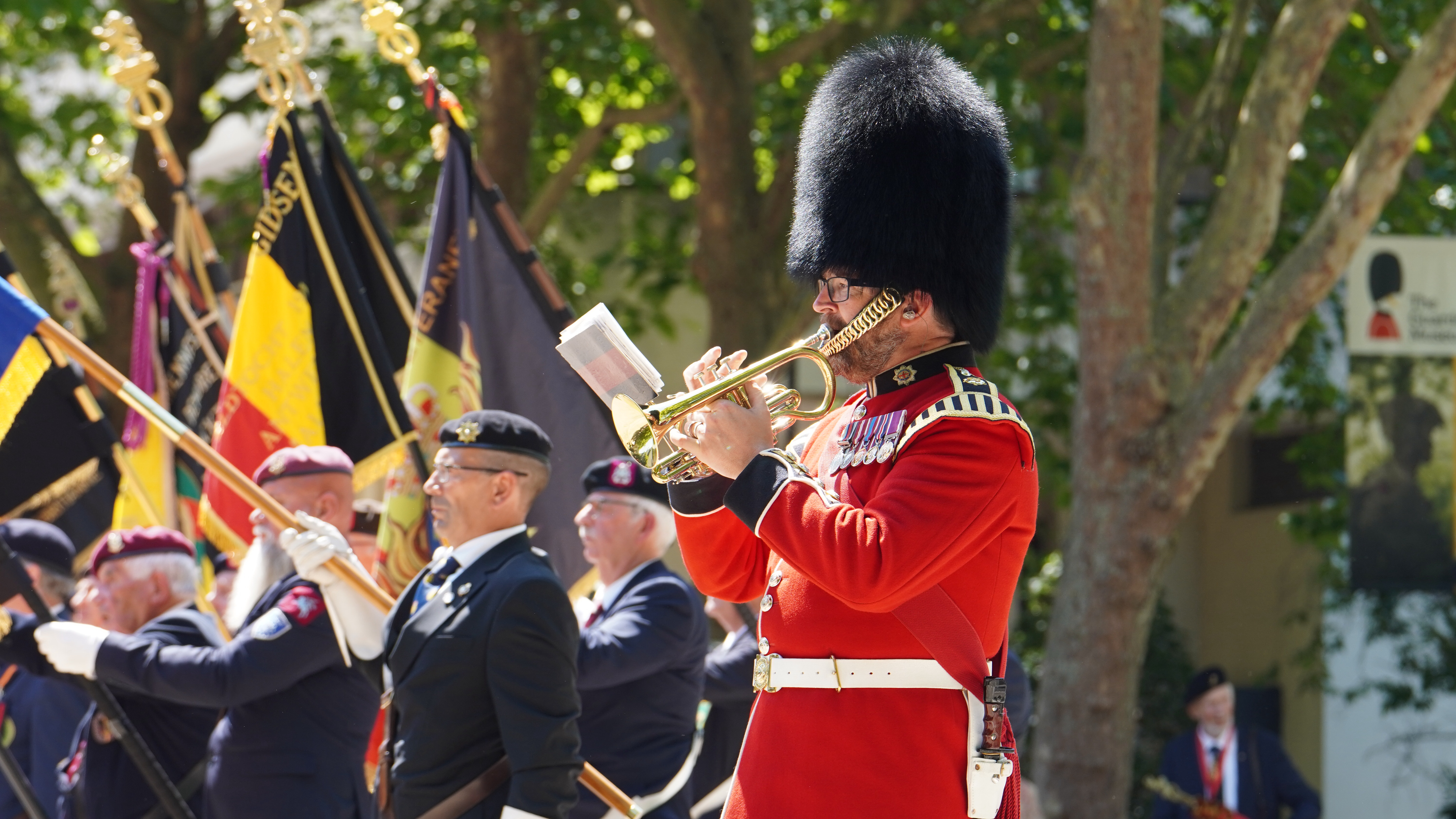 A parade features participants in formal military attire. A person in a red uniform and bearskin hat plays the trumpet. 