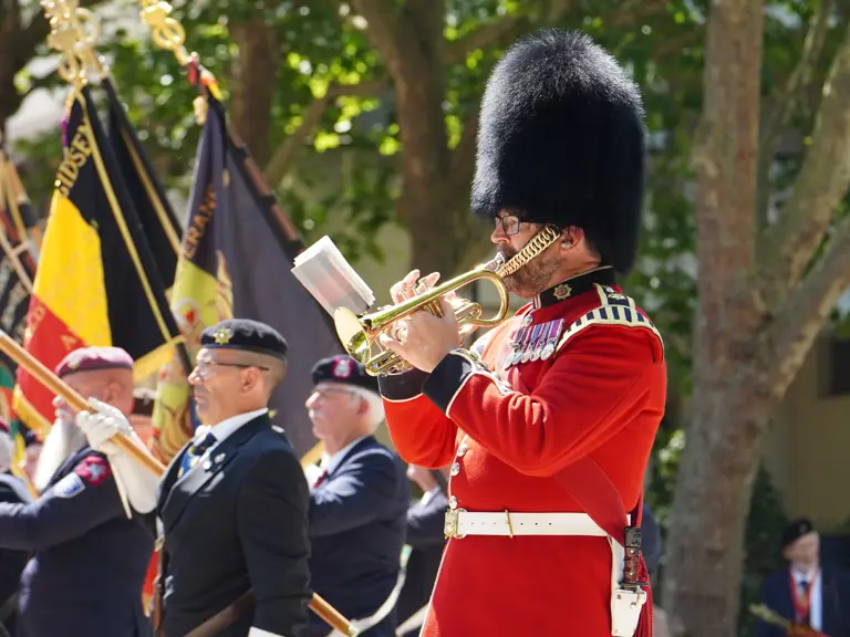 A parade features participants in formal military attire. A person in a red uniform and bearskin hat plays the trumpet.