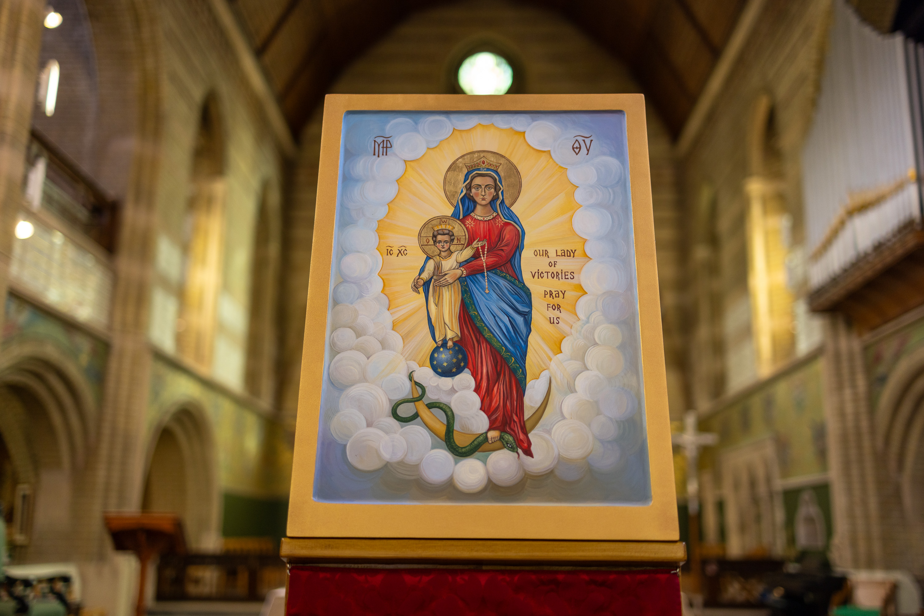 Icon of Our Lady of Victories holding a child, surrounded by clouds and radiant light inside a church.