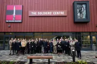 A large group of people stand outside of a red building, some are wearing formal civilian attire and some are wearing military uniform.