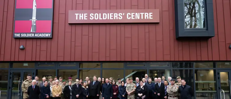 A large group of people stand outside of a red building, some are wearing formal civilian attire and some are wearing military uniform.