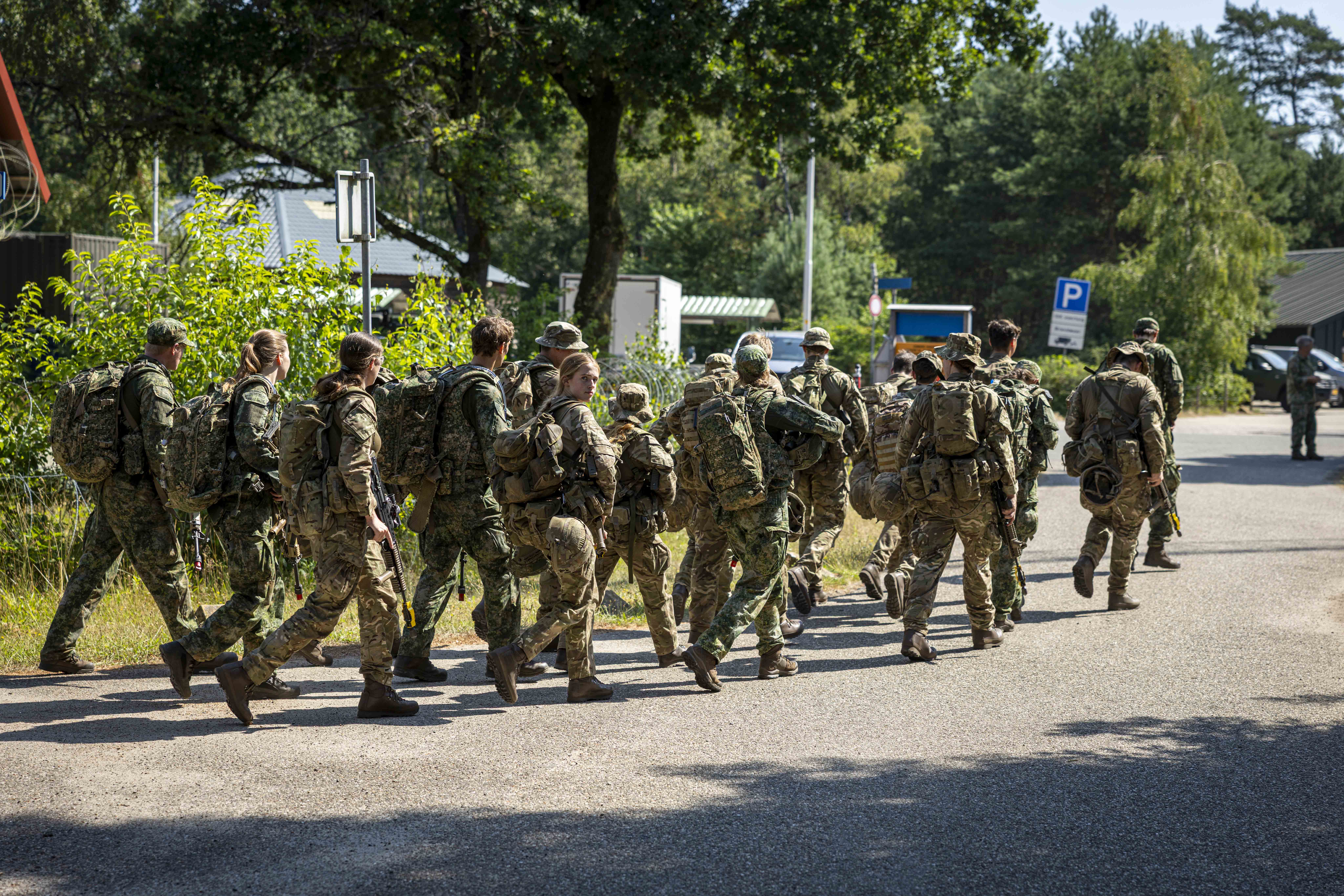 A group of soldiers in camouflage uniforms and backpacks are walking along a road in a wooded area. One soldier bears a German flag patch. Trees and parked cars are visible in the background.