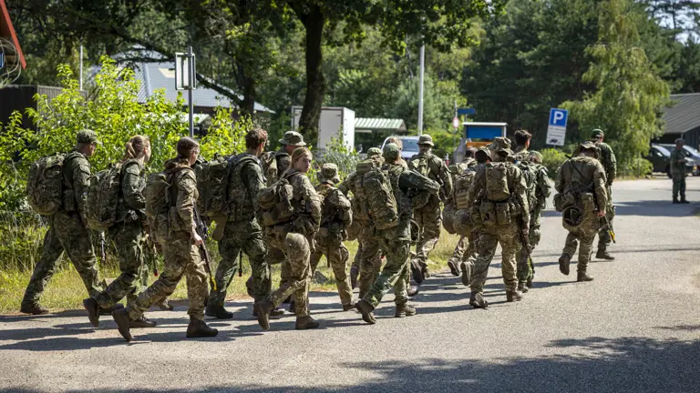 A group of soldiers in camouflage uniforms and backpacks are walking along a road in a wooded area. One soldier bears a German flag patch. Trees and parked cars are visible in the background.