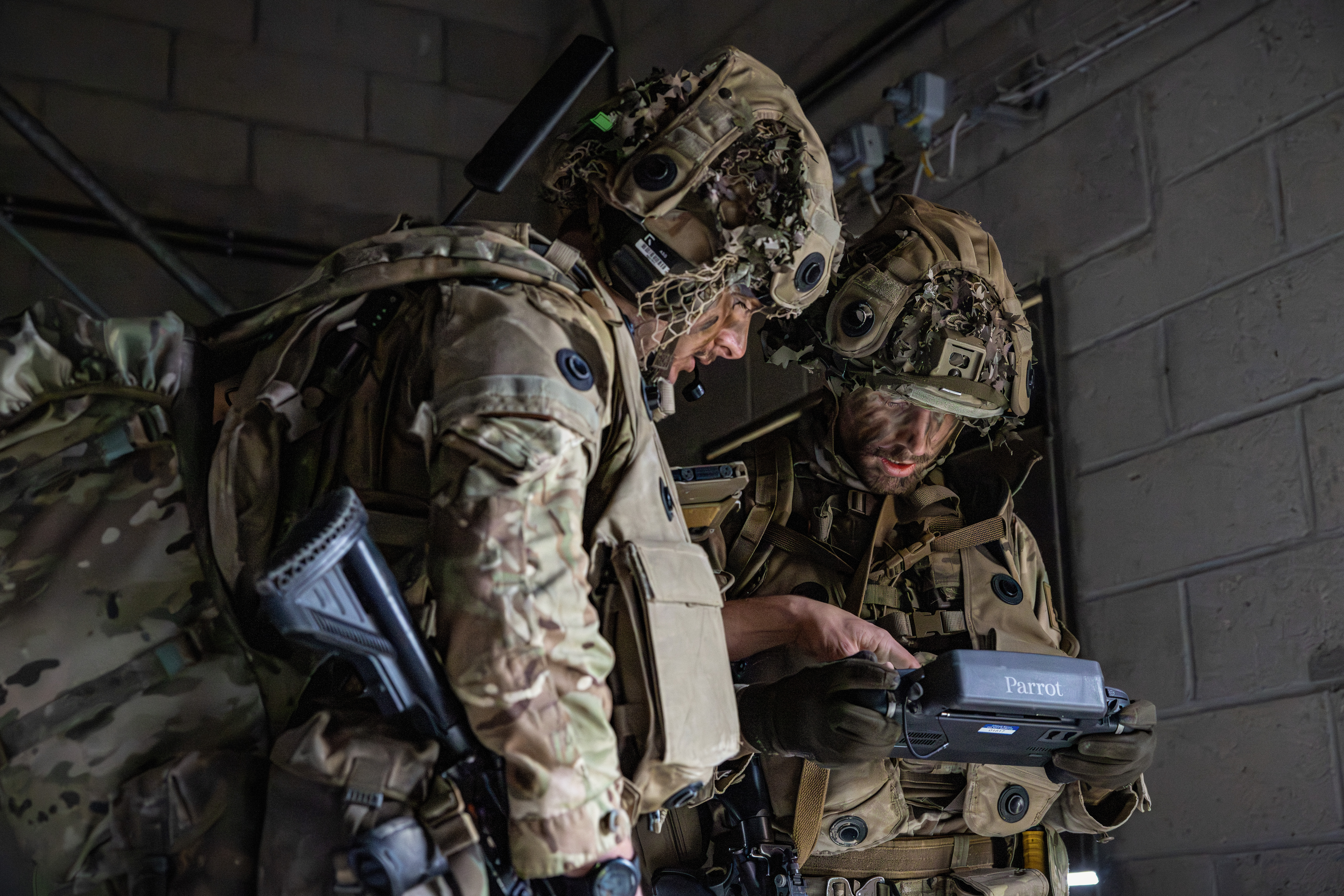 Two individuals in camouflage military uniforms and helmets are indoors, closely examining a device labeled 