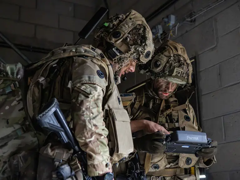 Two individuals in camouflage military uniforms and helmets are indoors, closely examining a device labeled
