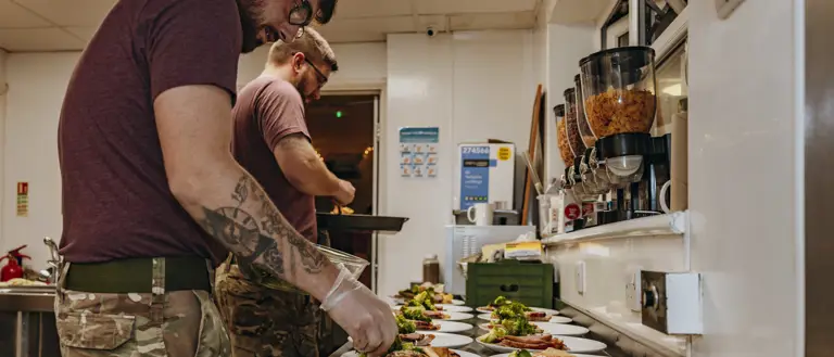 Two men in purple t-shirts and camo trousers prepare over a dozen plates of traditional Christmas food.