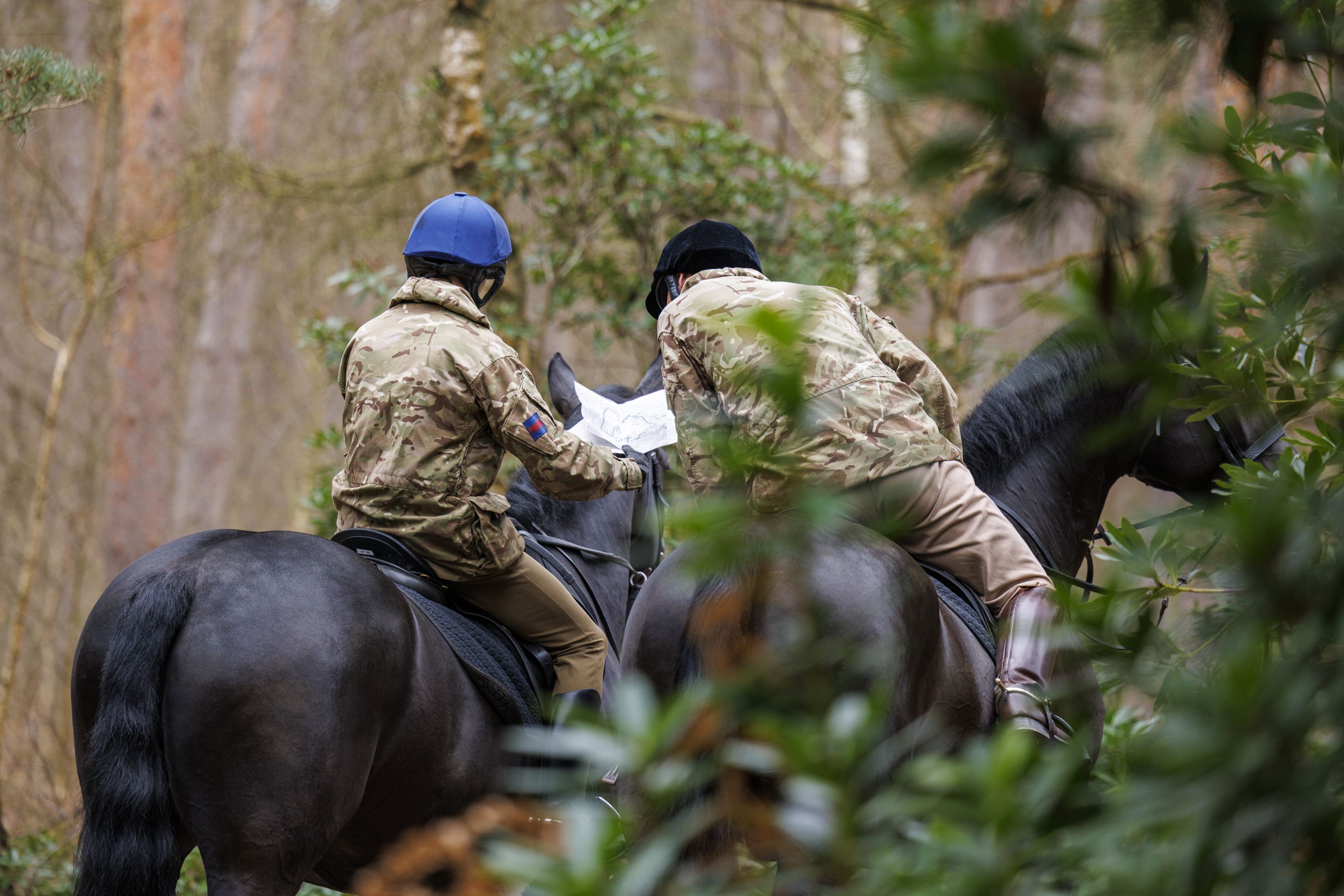 Two individuals in camouflage jackets and helmets, riding horses through a forest, are looking at a map in their hands. 