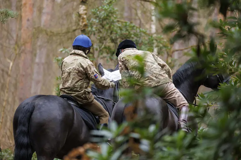 Two individuals in camouflage jackets and helmets, riding horses through a forest, are looking at a map in their hands.