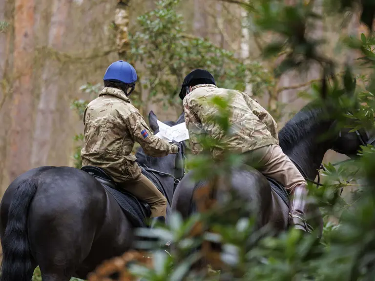 Two individuals in camouflage jackets and helmets, riding horses through a forest, are looking at a map in their hands.