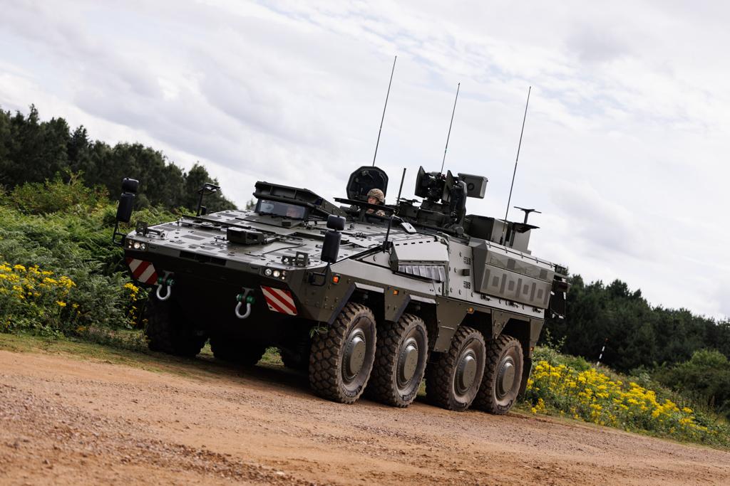 An armoured military vehicle, a Boxer, is on a dirt path surrounded by greenery and yellow flowers. 