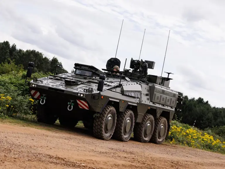 An armoured military vehicle, a Boxer, is on a dirt path surrounded by greenery and yellow flowers.