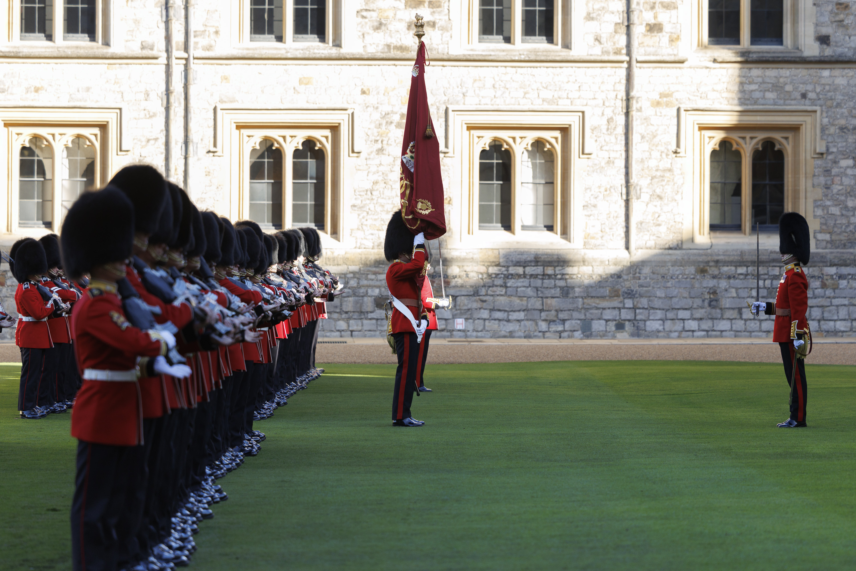 British guards in red uniforms and bearskin hats standing in formation on a lawn outside of a historic stone building.