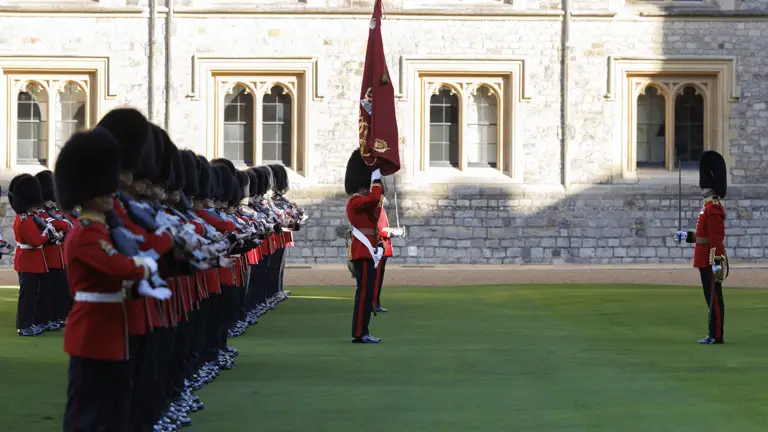 British guards in red uniforms and bearskin hats standing in formation on a lawn outside of a historic stone building.