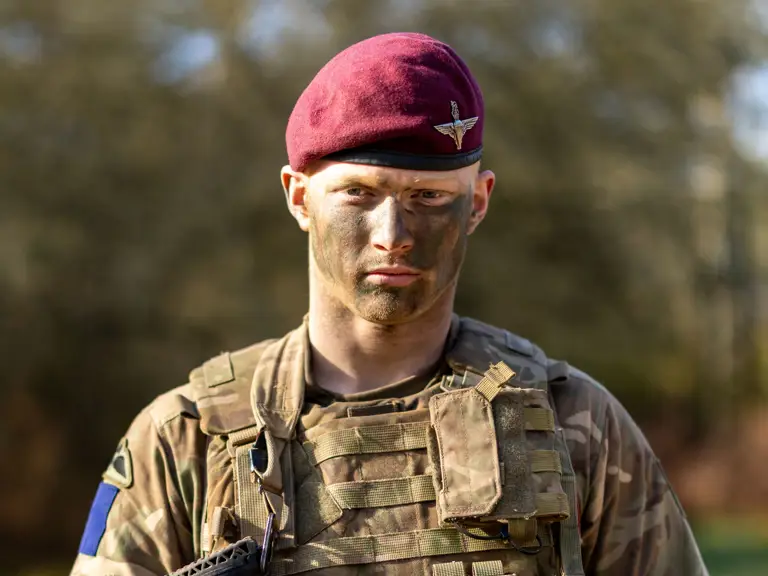 The image shows a person in military camouflage attire, wearing a maroon beret with an emblem.